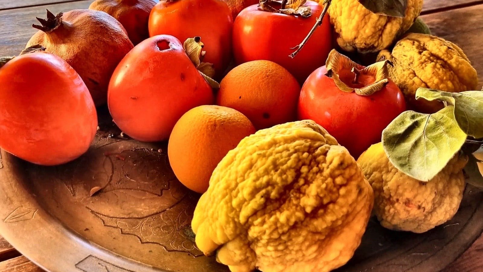 Bļoda ar hurmām, granātāboliem un citroniem uz koka galda
